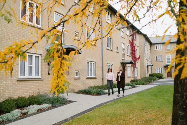 Image of 2 people walking outside of student accommodation with tree in forefront of image.